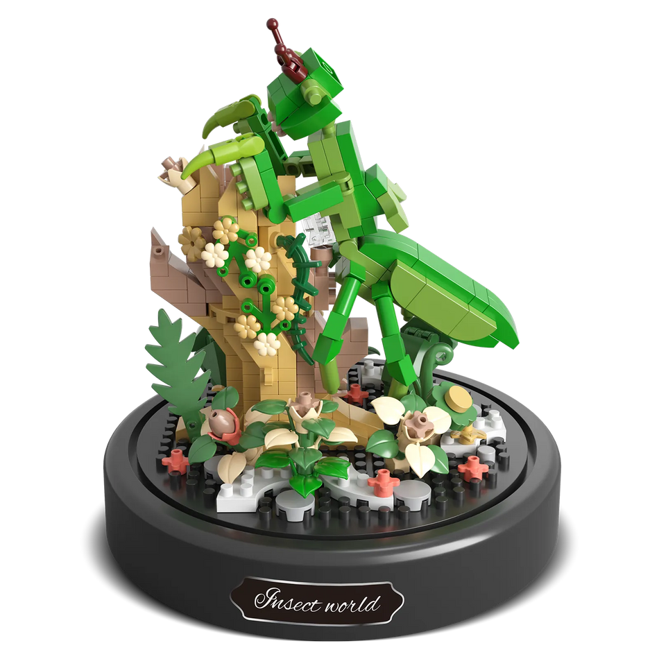 Produktfoto der Balody Miniblocks Gottesanbeterin auf einem Zweig mit Blumen und pflanzlichem Boden, unter einer Glocke