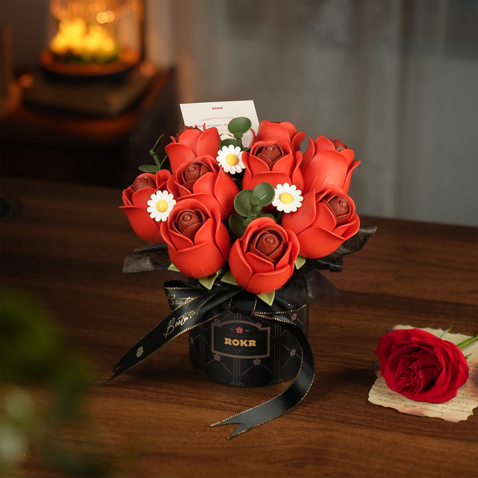 Atmosphärische Präsentation der Robotime Red Rose Bouquet, Kunststoff-Bausatz für einen Strauß roter Rosen und weißer Gänseblümchen in einer schwarzen Vase mit schwarzem Band darum